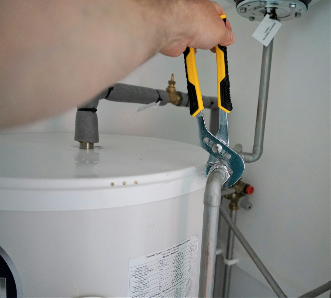 A person uses channel lock pliers to tighten a metal fitting on a water heater, indicating a plumbing maintenance task.