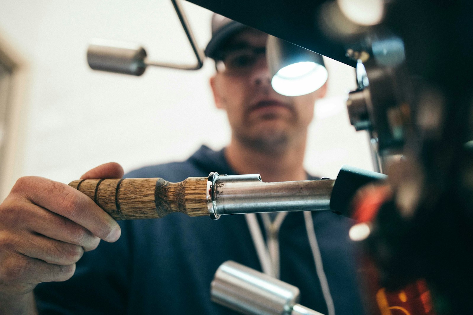 A man carefully works on a machine, holding a tool with a wooden handle, focused on a metallic component under a bright light.