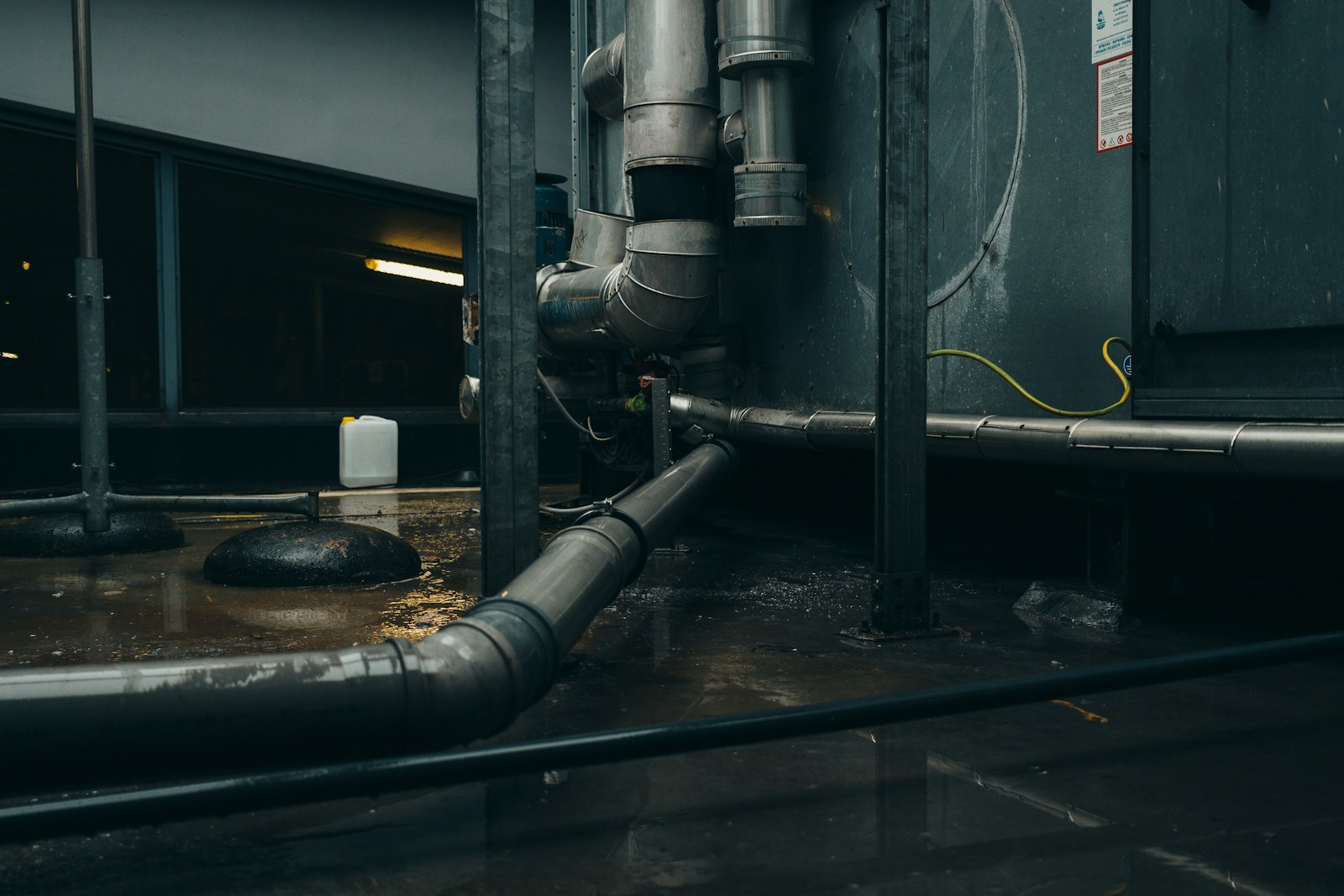 Industrial pipes and equipment in a dimly lit space, with water pooling on the floor. The image highlights maintenance needs in facility management.